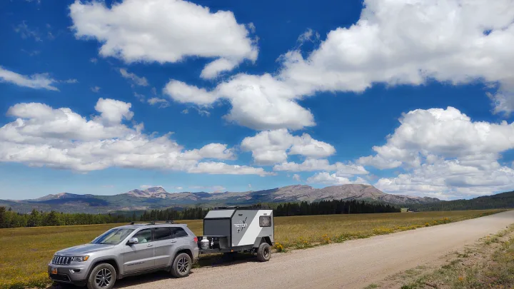 Off-grid RV rental adventure! This modern camper, towed by a Jeep, is parked on a dirt road near mountains.. TetonX Porter 2022