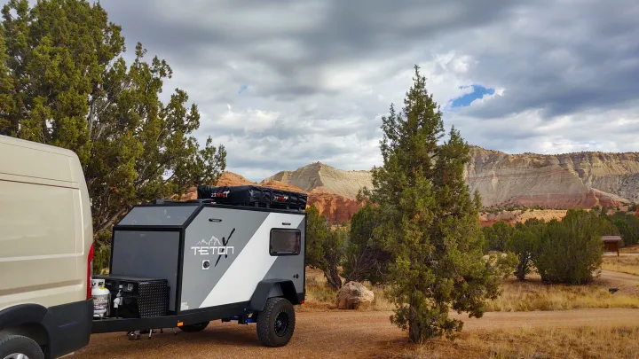An RV rental towing an off-road adventure trailer is parked at a scenic desert campsite with layered rock cliffs.. TetonX Porter 2022