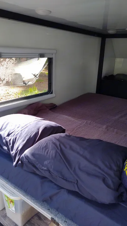 Inside this RV rental: A cozy sleeping space with a bed, blue bedding, purple blanket, window, and floor storage.. TetonX Porter 2022