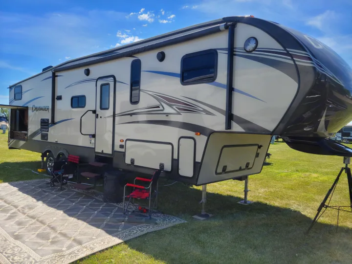Spacious fifth-wheel RV rental set up at a green campground on a sunny day, with an outdoor mat and chairs.. Crusader Crusader Fifth Wheel 2016