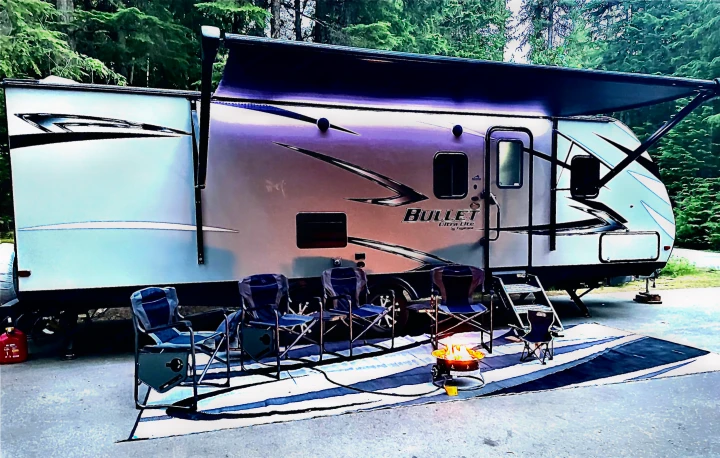 A modern RV rental campsite setup, featuring an extended awning, outdoor rug, chairs, and a glowing portable fire pit.. Keystone Bullet 2020