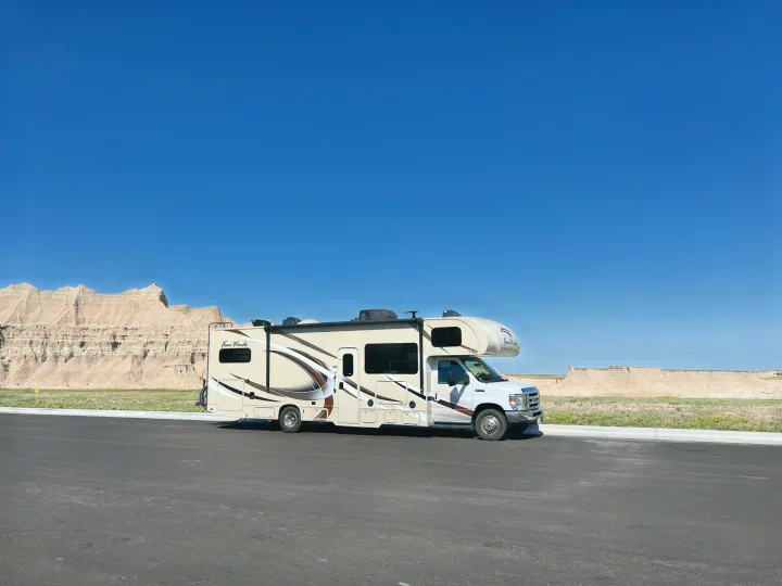 A beige and white RV rental is parked on a paved lot, with dramatic light-colored Badlands formations under a clear blue sky.. Thor Motor Coach Four Winds 2018