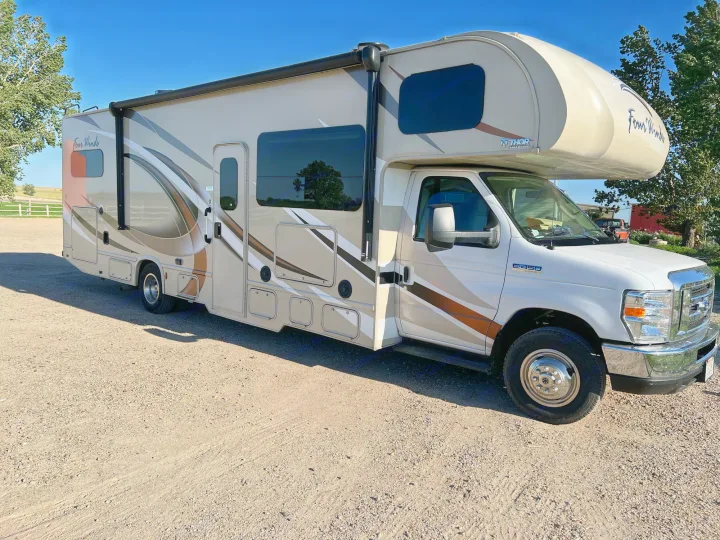 This beige Thor Four Winds Class C motorhome, ideal for an RV rental adventure, is parked on gravel under a clear sky.. Thor Motor Coach Four Winds 2018
