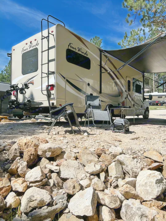 A tan rv rental is set up at a sunny campsite with an extended awning, chairs, and a grill on rocky ground.. Thor Motor Coach Four Winds 2018