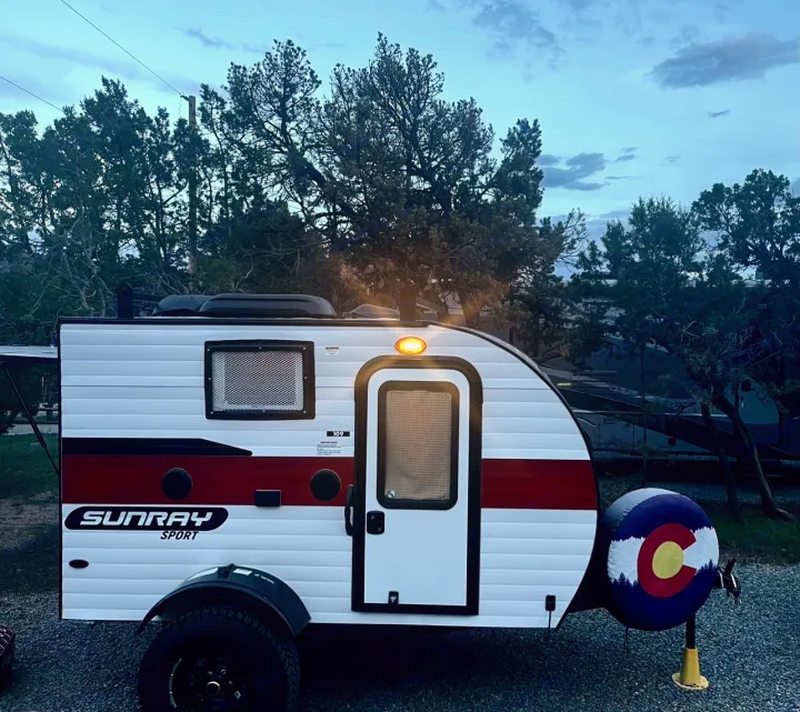 This compact Sunray Sport travel trailer, white with red stripes, is parked at a campsite. Ideal for your next RV rental adventure.. Sunset Park RV SunRay 2020