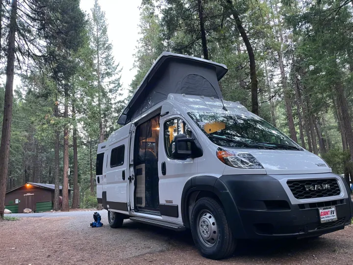 White pop-top rv rental van parked at a forest campsite. Side door open; light illuminates the interior.. Winnebago Winnebago 2023