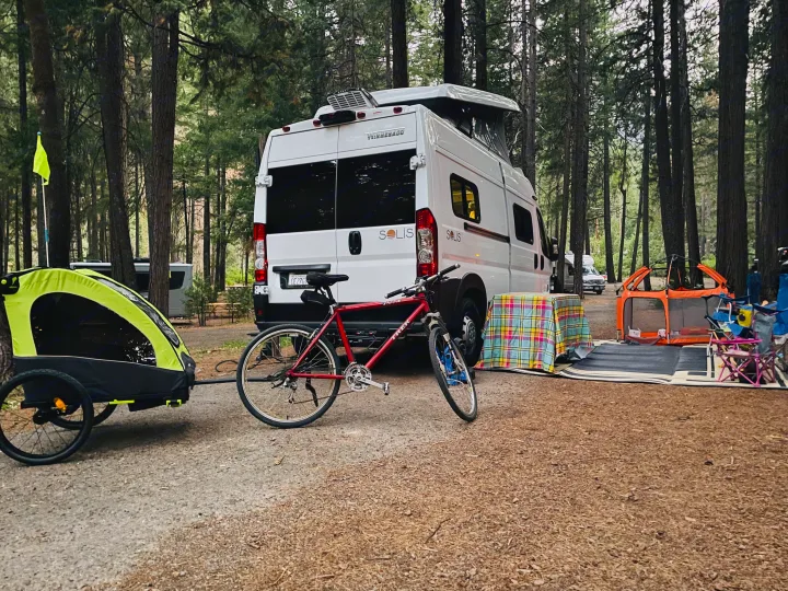 Camping adventure with an RV rental: A white camper van, bike, trailer, and outdoor gear set up in a lush forest campground.. Winnebago Winnebago 2023