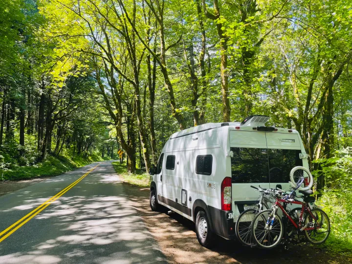 White RV rental van with bikes on the back, parked on a scenic, tree-lined road with dappled sunlight. Perfect nature escape.. Winnebago Winnebago 2023