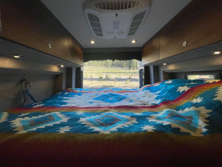 Experience comfort in this RV rental: a plush bed with a colorful blanket and serene forest views.. Storyteller Overland Storyteller Overland Class B 2021