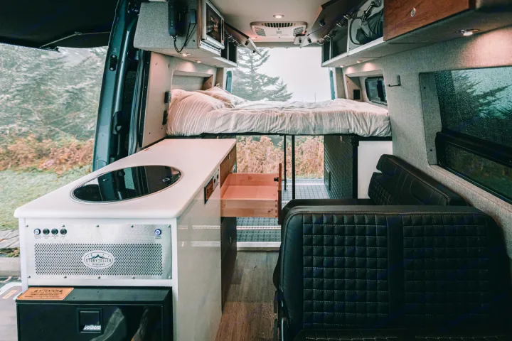 Modern RV rental interior with an elevated bed, compact kitchenette, and black quilted seating. Rear doors reveal a nature view.. Storyteller Overland Storyteller Overland Class B 2021