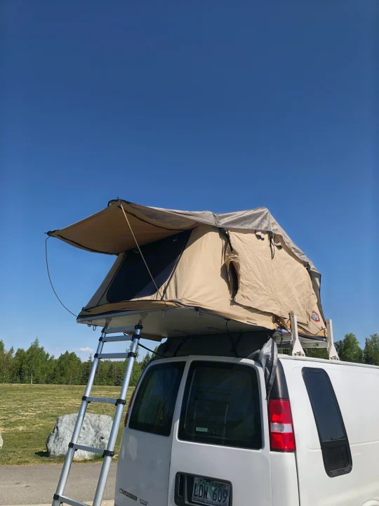 This RV rental features a white van with a deployed tan rooftop tent and ladder, ready for outdoor adventure under a clear blue sky.. Chevrolet Other 2006