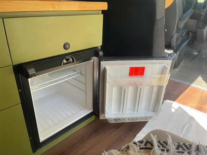 Open mini-fridge in an RV rental's olive-green camper van interior, showing clean shelves and door storage.. Freightliner Sprinter 2010
