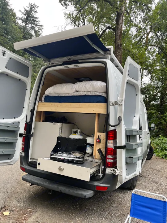 An RV rental camper van, rear open, reveals a compact bed, pull-out kitchen, and extended side awning.. Freightliner Sprinter 2010