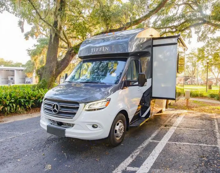 White Tiffin RV rental, a Mercedes-Benz Sprinter motorhome, with its slide-out extended in a park setting.. Tiffin Motorhomes Wayfarer LW 2023