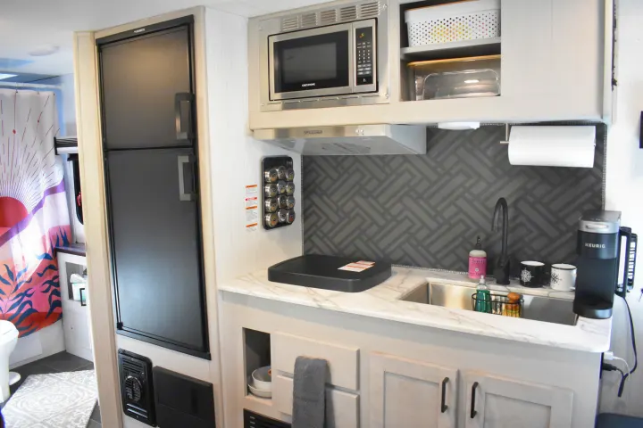 Compact rv rental kitchen: black fridge, microwave, sink, coffee maker, geometric backsplash. Bathroom area also visible.. Heartland RVs Mallard 2022