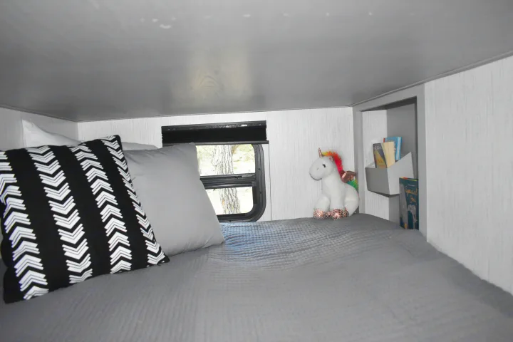 Cozy RV rental bunk bed featuring a grey blanket, decorative pillow, window, and a playful unicorn toy.. Heartland RVs Mallard 2022