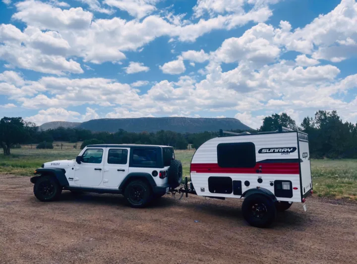Adventure-ready rv rental: White Jeep Willys towing a Sunray Sport camper on a dirt road, mountains & clouds.. Sunset Park RV SunRay 2020
