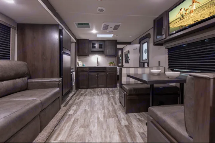Modern RV rental interior: dark wood kitchen, cozy sofa, dining booth, and mounted TV with beach scene.. Grand Design Transcend Xplor 2022