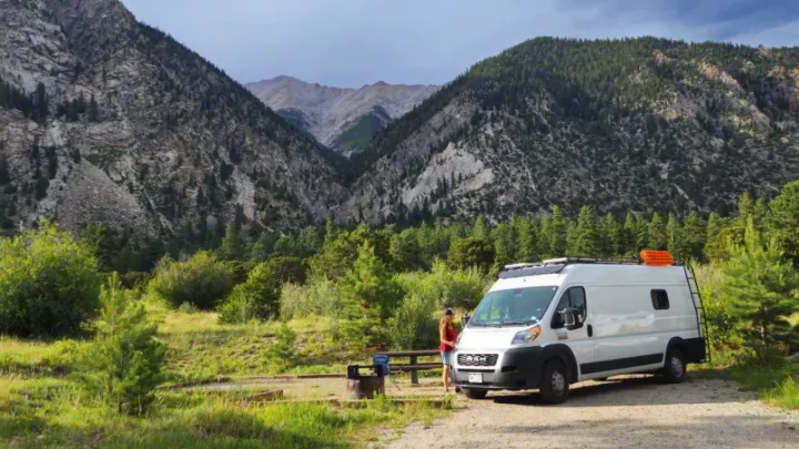 RV rental van at a scenic mountain campsite. A person stands by a white camper van, picnic table, and fire pit.. Other Other 2021