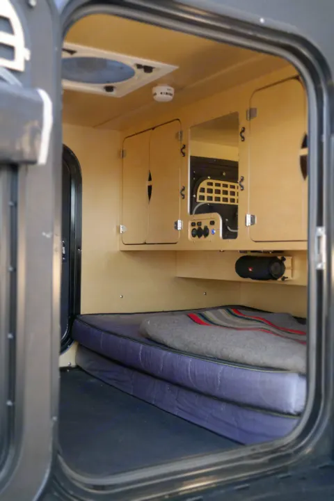 View inside a compact RV rental: wood cabinets, control panel, mattress with blanket, ceiling fan, and speaker.. Oregon Trail'R TerraDrop ALPHA 2022