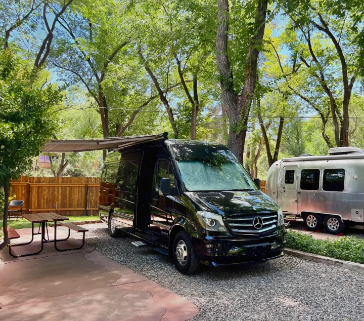 Black Mercedes Sprinter **RV rental** parked in a shaded campsite with an Airstream trailer and picnic table.. Airstream Interstate 2016