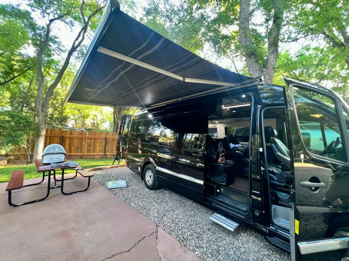 Shady RV rental spot: A black camper van with extended awning is parked next to a picnic table with a grill on a concrete patio.. Airstream Interstate 2016