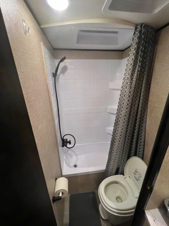 RV rental's compact bathroom features a white shower, black fixtures, and toilet, with a gray patterned shower curtain.. Jayco Jay Flight SLX 2022