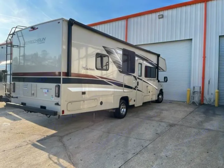 A beige Coachmen Leprechaun RV rental is parked, showing its rear ladder & extended awning, ready for travel.. COACHMEN LEPRECHAUN 2021