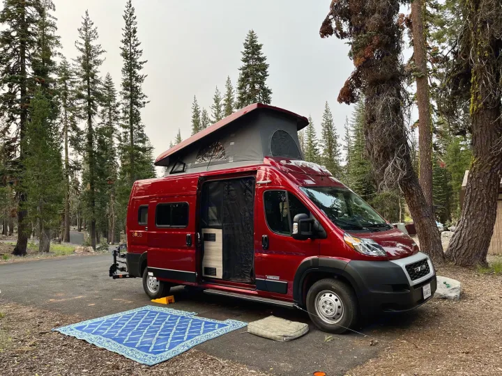 This vibrant red Winnebago Solis rv rental is parked at a forest campsite, pop-top extended, with an open door.. Winnebago Solis 2021