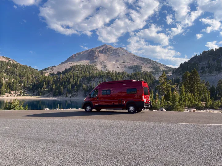 Red RV rental parked by a calm mountain lake with a majestic peak, surrounded by pines under a blue sky.. Winnebago Solis 2021