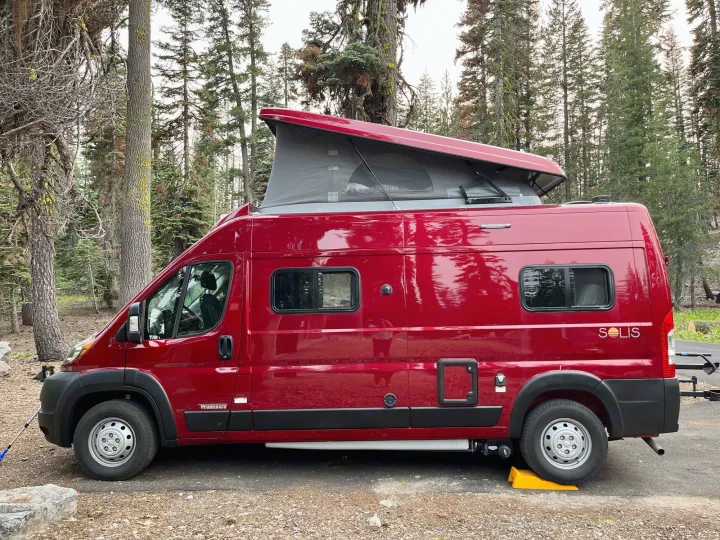 Red Winnebago Solis RV rental with pop-top raised, parked in a forest campsite, leveled with a yellow block.. Winnebago Solis 2021