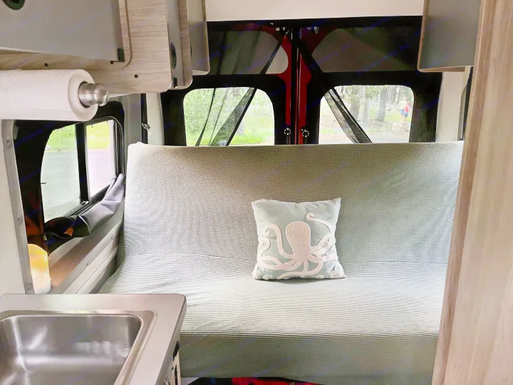 Inside a comfortable rv rental: Kitchenette, striped seating/bed, and a view of the outdoors with trees and people.. Winnebago Solis 2021
