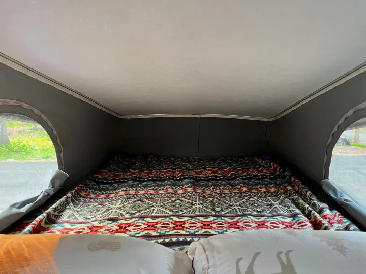 Cozy RV rental pop-top bed with vibrant patterned blanket. Windows show lush greenery outside.. Winnebago Solis 2021