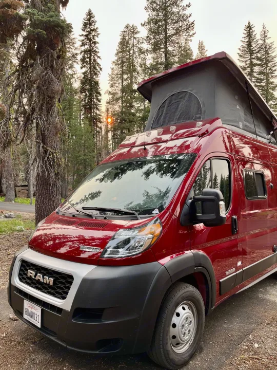 Red Winnebago pop-top camper van, an RV rental, parked in a pine forest with a visible sunset.. Winnebago Solis 2021