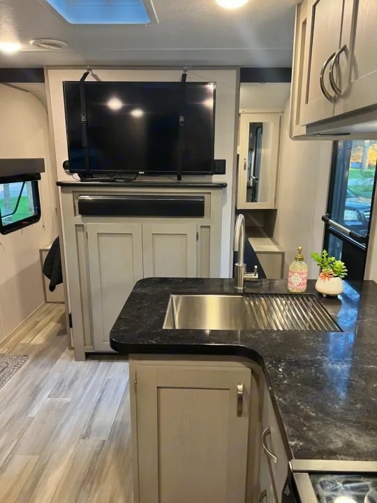 Modern rv rental interior: kitchen with black countertops, stainless sink, and wall-mounted TV. Features light cabinetry and wood-look flooring.. PrimeTime 31BHD 2021