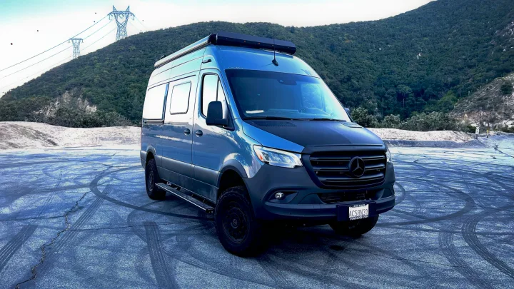 This rugged blue Mercedes-Benz Sprinter camper van, an ideal rv rental, is ready for adventure, parked before a forested mountain.. Winnebago Revel 2022
