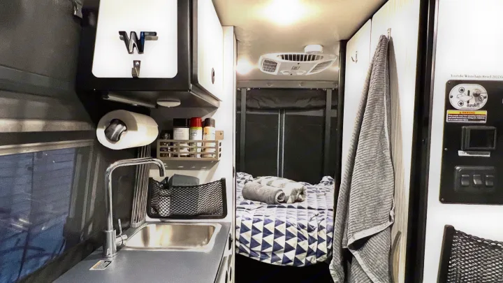 Cozy RV rental interior shows a compact kitchen with sink, paper towel holder, spices, and a geometric-patterned bed.. Winnebago Revel 2022