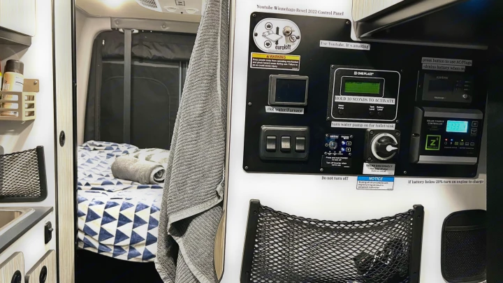 RV rental interior: A camper van's control panel, showing switches and displays for heating, water, and solar charge, beside a cozy bed.. Winnebago Revel 2022