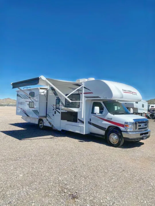White Class C RV rental with extended awnings and slide-out, parked on a gravel lot under a clear sky.. Thor Freedom Elite 2020