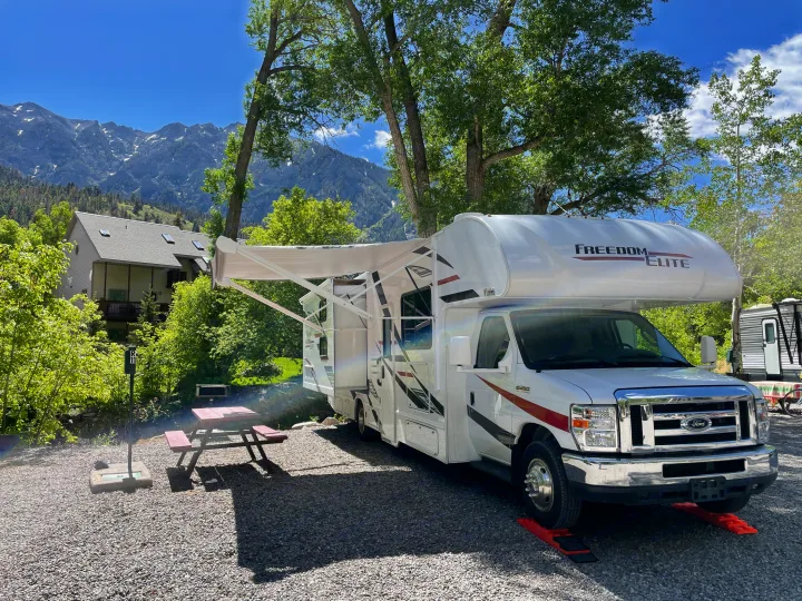 White RV rental parked at a scenic mountain campsite. Awning extended, picnic table beside. Trees & house visible.. Thor Freedom Elite 2020