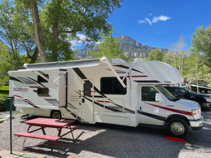 A white RV rental is parked at a gravel campsite with a red picnic table, surrounded by lush trees and distant mountains.. Thor Freedom Elite 2020