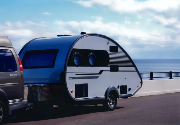 A sleek teardrop RV rental is hitched to a van, parked by a scenic coastal road with a blue sky and ocean view, ready for adventure.. Nucamp Tab 400 2022