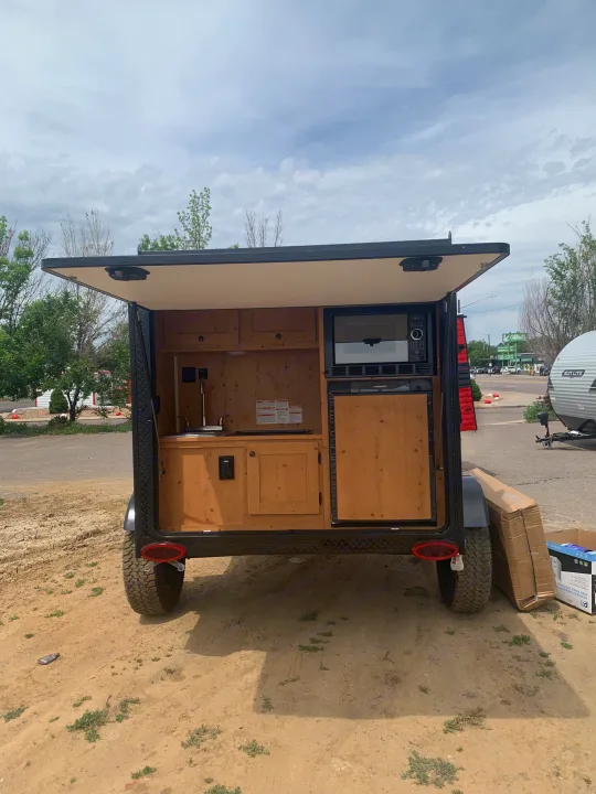 Compact RV rental with its rear open, revealing a built-in outdoor kitchen with microwave, fridge, and sink. Ready for travel!. Sunset Park RV SunRay 2021