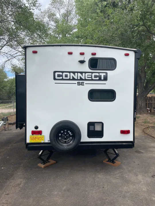 Rear view of a white RV rental, parked outdoors with a spare tire and stabilizing jacks extended.. Connect SE 2021