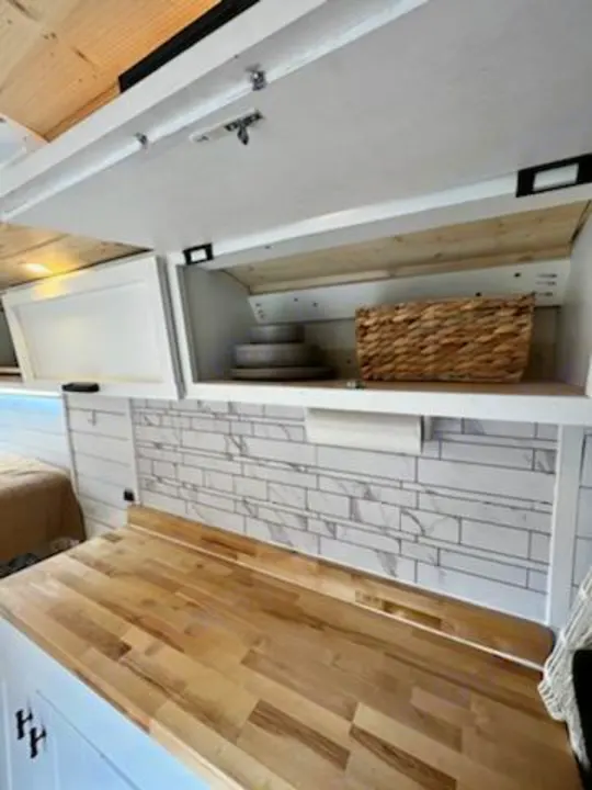 RV rental kitchen: Wood countertop, tile backsplash, & accessible overhead storage with dishes & a basket.. Dodge Ram 3500 Pro master 2022