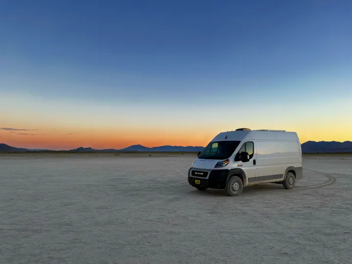 White RV rental van on a vast, cracked desert plain under a sunset sky, with distant mountains. Perfect for travel.. RAM ProMaster 2022