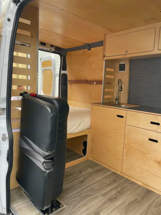 Clean, modern camper van interior for RV rental. Features wood paneling, kitchenette, bed, and rear seating.. RAM ProMaster 2022