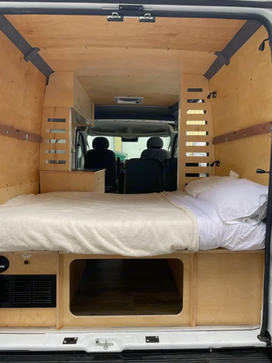 Interior of a comfortable, custom-built camper van, ideal for an RV rental. Features a cozy bed, wood paneling, and storage.. RAM ProMaster 2022