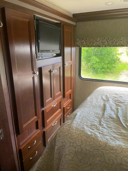 This RV rental bedroom features a dark wood cabinet with a built-in TV, drawers, and a cozy bed by a window.. Forest River Sunseeker 2017