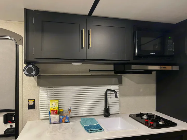 A compact RV rental kitchen featuring dark overhead cabinets, microwave, sink with black faucet, and two-burner stovetop.. Keystone Springdale 2022